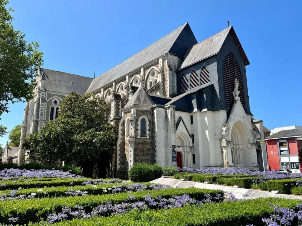 Photographie de la façade de l'église Saint-Nazaire avec au premier plan des parterres d'agapanthes en fleurs.