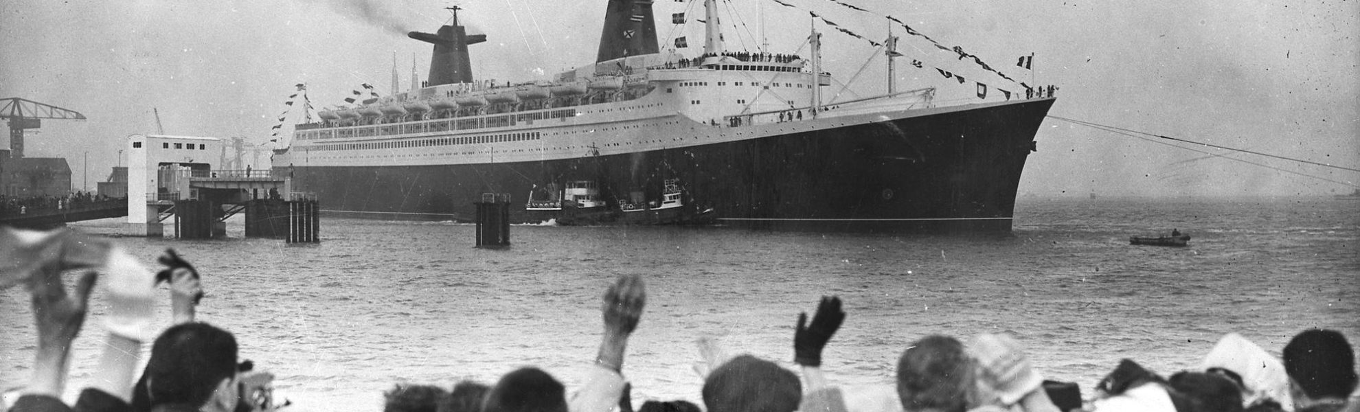 Photographie noir et blanc du paquebot France sortant du port de Saint-Nazaire, au premier plan une foule agite les bras.