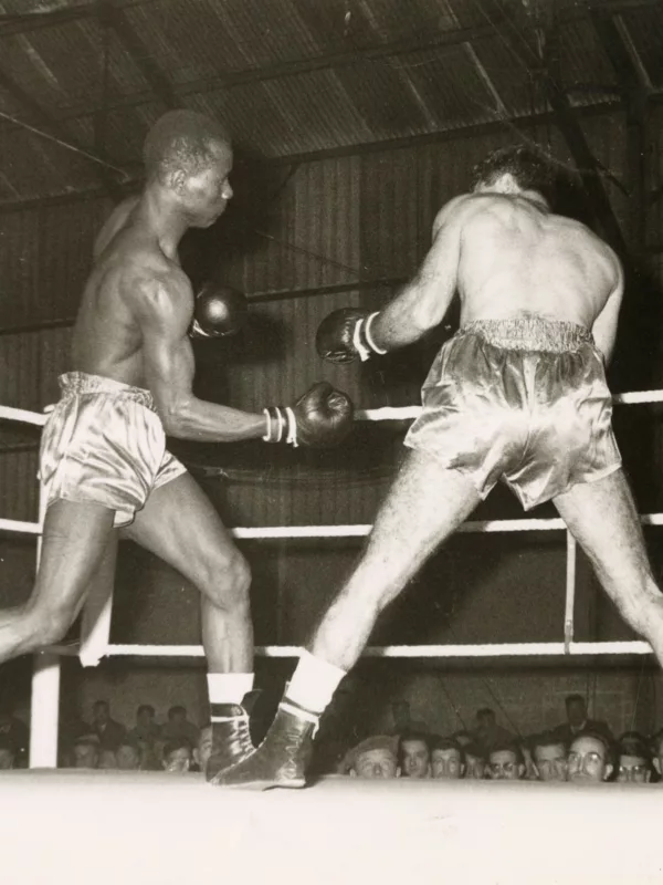 Combat de boxe sur un ring entre Souleymane Diallo, à gauche et à droite, dans les hangars de la chambre de commerce et d'industrie au Petit Maroc. Une foule qui regarde le combat est assise en arrière plan.