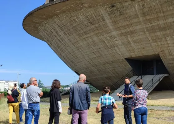 Photographie en couleurs d'un groupe de visiteurs et d'un guide devant la Soucoupe, le palais des sports de Saint-Nazaire.