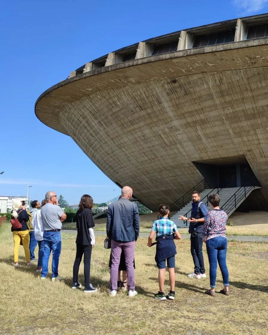 Photographie en couleurs d'un groupe de visiteurs et d'un guide devant la Soucoupe, le palais des sports de Saint-Nazaire.