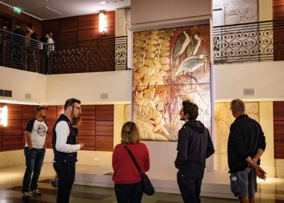 Un médiateur parle à un groupe de visiteurs devant un décor en laque provenant du paquebot Normandie (1935) et représentant des renards et des marabouts.