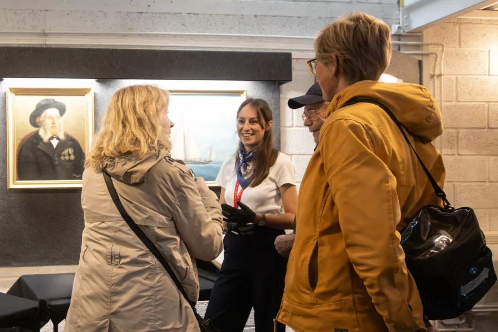 Médiatrice lors d'une visite-flash à l'Écomusée de Saint-Nazaire souriant à des visiteurs devant un tableau représentant un pilote de Loire.