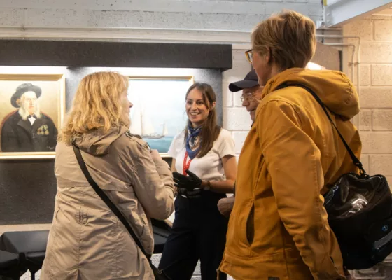 Médiatrice lors d'une visite-flash à l'Écomusée de Saint-Nazaire souriant à des visiteurs devant un tableau représentant un pilote de Loire.