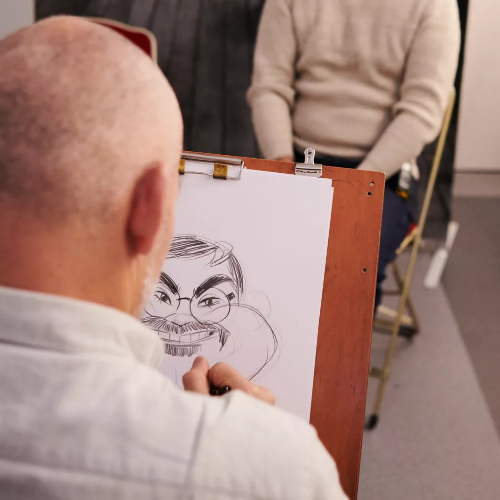 Photographie prise au-dessus de l'épaule d'un dessinateur caricaturiste alors qu'il dessine le visage d'un homme.