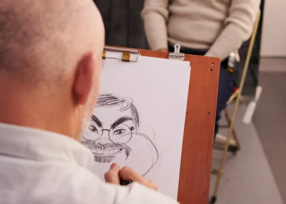 Photographie prise au-dessus de l'épaule d'un dessinateur caricaturiste alors qu'il dessine le visage d'un homme.